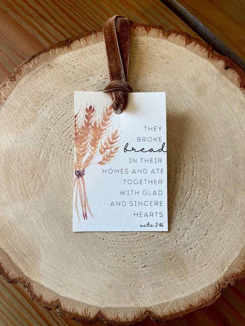 K&ouml;nnte beinhalten: Ein rechteckiges Etikett mit einem braunen Samtband, das eine Aquarellillustration von Weizen und den Text "They broke bread in their homes and ate together with glad and sincere hearts acts 2:46." zeigt.