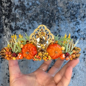 May include: A decorative crown featuring a central gold filigree arch with a large amber-colored crystal. The base is adorned with two orange pumpkins, small yellow and orange berries, green foliage, pinecones, and tiny red heart-shaped beads.