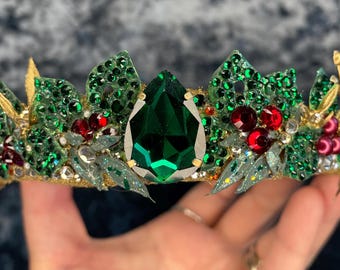 May include: A green and gold crown with a large emerald green gemstone in the center. The crown is decorated with red and green rhinestones and gold accents.