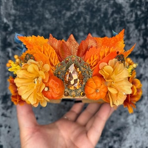 May include: A decorative headband featuring an arrangement of orange and yellow fabric flowers, artificial autumn leaves, and small orange pumpkin figures. A central bronze-toned filigree setting holds a large, faceted amber-colored teardrop gem.