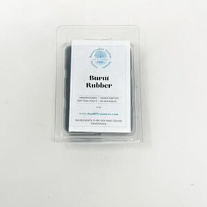May include: A package of soy wax melts in a clear plastic container. The label reads "Burnt Rubber" with additional text. The wax is dark gray. The product is hand-poured and made with pure soy wax.