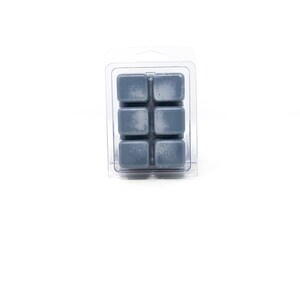 May include: A clear plastic clamshell package containing six gray wax melt cubes. The cubes are arranged in two rows of three. The wax has a slightly textured appearance. The background is plain white.
