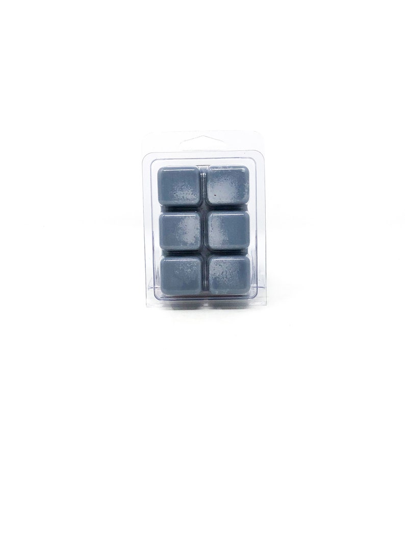May include: A clear plastic clamshell package containing six gray wax melt cubes. The cubes are arranged in two rows of three. The wax has a slightly textured appearance. The background is plain white.