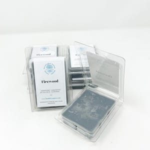 May include: Several packages of soy wax melts in clear plastic clamshells. The label reads "Firewood" with additional text. The wax melts are a dark gray color. The product is hand-poured and handcrafted.