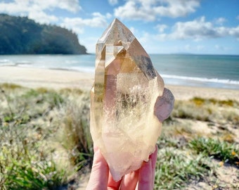 May include: A large, translucent quartz crystal with a pointed tip and a light brown hue. The crystal is held up against a backdrop of a beach, ocean, and blue sky.