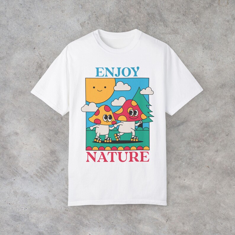 A white Enjoy Nature t-shirt lays flat, featuring a playful design of mushrooms holding hands and rollerblading under the sun. The cheerful, nature-inspired graphic evokes a sense of fun and positivity, perfect for mushroom lovers.