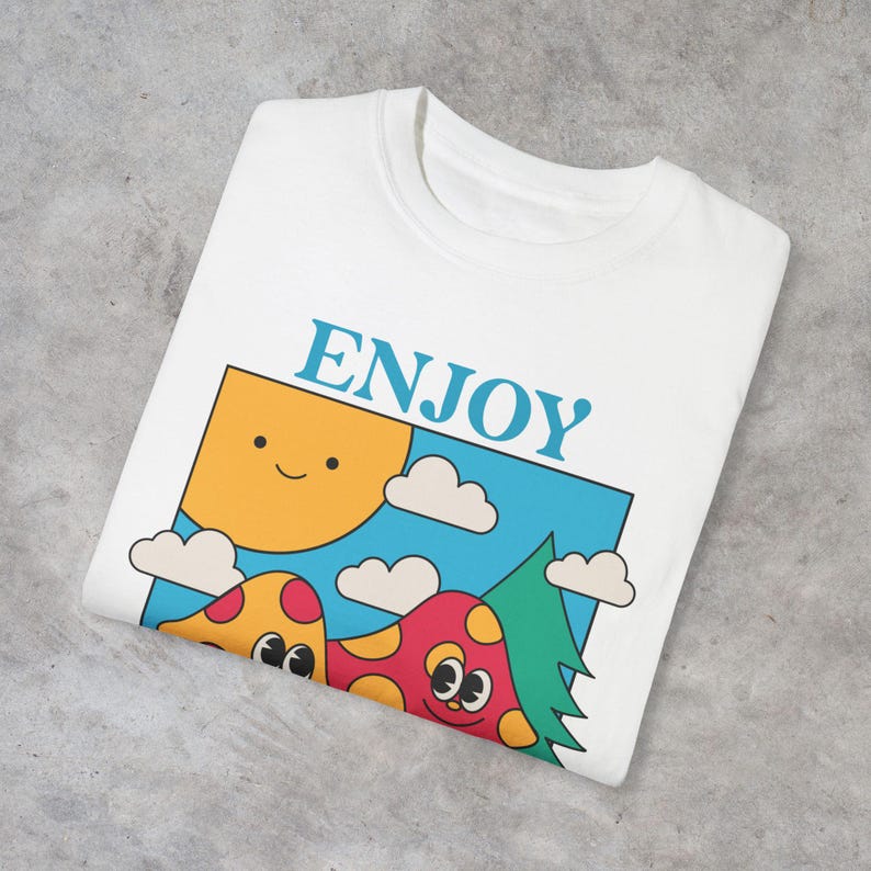 A white Enjoy Nature t-shirt lays folded, featuring a playful design of mushrooms holding hands and rollerblading under the sun. The cheerful, nature-inspired graphic evokes a sense of fun and positivity, perfect for mushroom lovers.