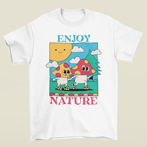A white Enjoy Nature t-shirt lays flat, featuring a playful design of mushrooms holding hands and rollerblading under the sun. The cheerful, nature-inspired graphic evokes a sense of fun and positivity, perfect for mushroom lovers.