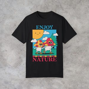 A black Enjoy Nature t-shirt lays flat, featuring a playful design of mushrooms holding hands and rollerblading under the sun. The cheerful, nature-inspired graphic evokes a sense of fun and positivity, perfect for mushroom lovers.