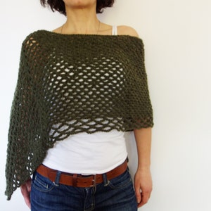 May include: Olive green crochet poncho with an open weave design. The poncho is draped over a white top and blue jeans. A brown leather belt with a silver buckle is worn at the waist. The poncho has a relaxed, off-the-shoulder fit.