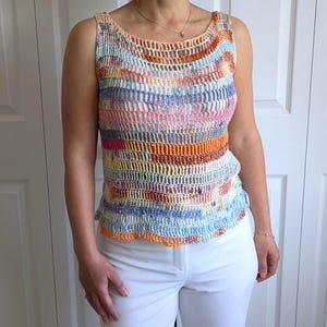 May include: A sleeveless, multi-coloured crochet top. The top features horizontal stripes of various colours, including pink, orange, blue, and beige. The crochet design has a textured, open weave. The top is paired with white trousers.