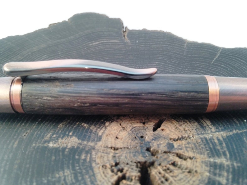 May include: A wooden pen with a copper band and a silver clip. The pen is resting on a wooden surface.