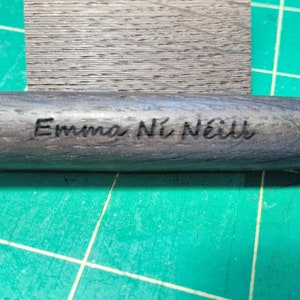 May include: A dark gray wooden cylinder with the name "Emma N&iacute; N&eacute;ill" engraved in black lettering.