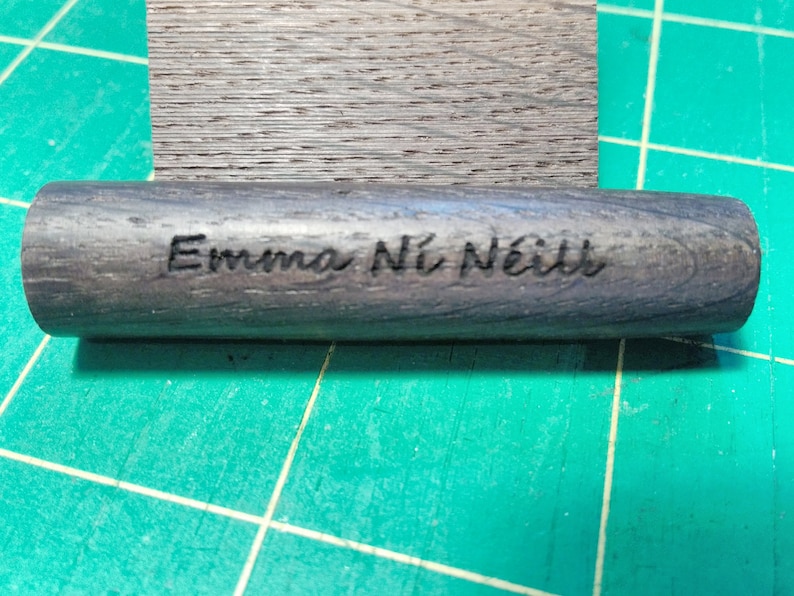 May include: A dark gray wooden cylinder with the name "Emma N&iacute; N&eacute;ill" engraved in black lettering.