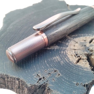 May include: A wooden pen with a copper-colored metal tip and a silver clip. The pen is resting on a dark wood surface.