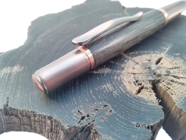 May include: A wooden pen with a copper-colored metal tip and a silver clip. The pen is resting on a dark wood surface.