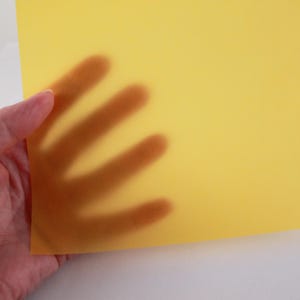 以下が含まれることがあります： 半透明の黄色い紙を手に持っています。手の影が紙を通して見え、光と影のコントラストを作り出しています。紙は滑らかでマットな仕上がりです。