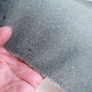 May include: A piece of blue-green suede fabric with a textured surface. The fabric is held by a hand, showing the rough, unfinished edge and small holes. The suede has a soft, matte appearance.