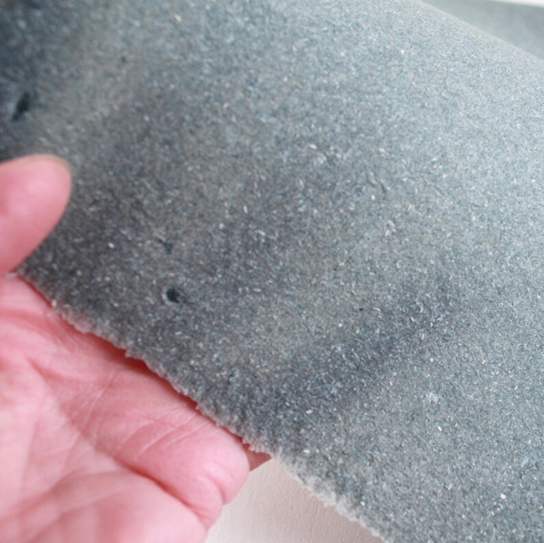 May include: A piece of blue-green suede fabric with a textured surface. The fabric is held by a hand, showing the rough, unfinished edge and small holes. The suede has a soft, matte appearance.