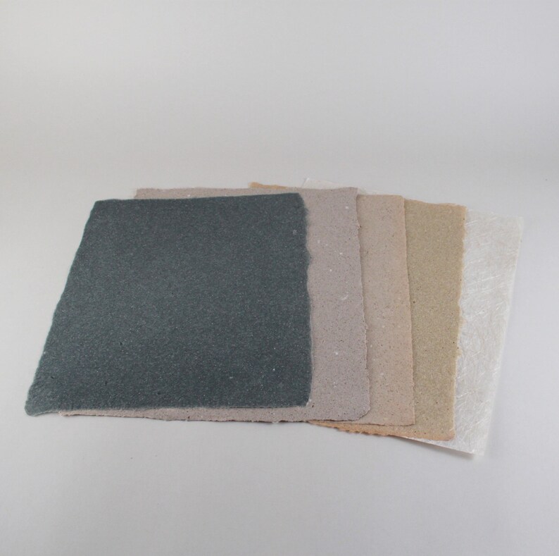 May include: A stack of handmade paper sheets in various neutral colors. The sheets include shades of gray, beige, and white, with a textured surface and deckled edges. The paper is arranged on a white background.