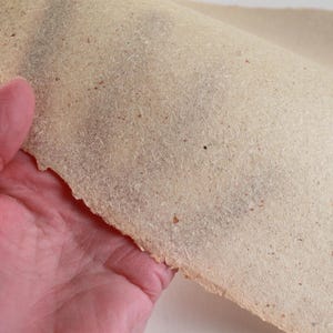 May include: A sheet of handmade paper with a rough, textured surface. The paper is a light beige color with small flecks of darker material throughout. The edge of the paper is uneven, suggesting it was torn by hand. The paper is being held up.