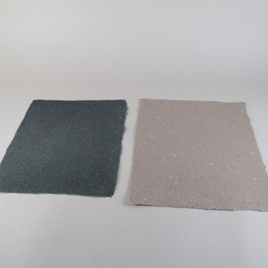 May include: Two rectangular sheets of handmade paper. One is a dark gray color, and the other is a light tan color. Both sheets have a rough, textured surface and irregular edges, suggesting a natural, artisanal quality.