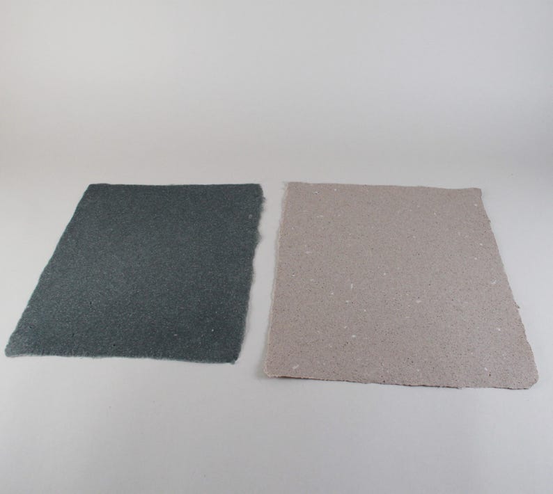 May include: Two rectangular sheets of handmade paper. One is a dark gray color, and the other is a light tan color. Both sheets have a rough, textured surface and irregular edges, suggesting a natural, artisanal quality.