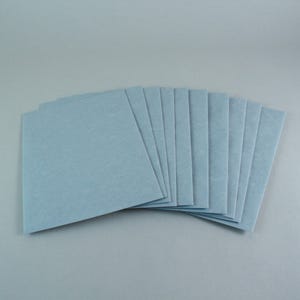 Könnte beinhalten: Ein Stapel hellblauer quadratischer Papierbögen, die auf einer grauen Oberfläche aufgefächert sind. Das Papier hat ein leicht strukturiertes Aussehen. Die Blätter sind in einem halbkreisförmigen Muster angeordnet, wobei die Kanten des Papiers sichtbar sind.