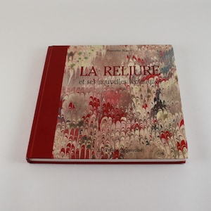 May include: A book titled "LA RELIURE et ses nouvelles techniques" with a marbled cover in red, white, and brown. The spine is a solid red colour. The book is lying on a white surface.