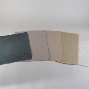 May include: A collection of handmade paper sheets in various neutral tones. The sheets range in color from dark gray to off-white, showcasing different textures and finishes. The edges of the paper are rough-cut, adding to the artisanal aesthetic.