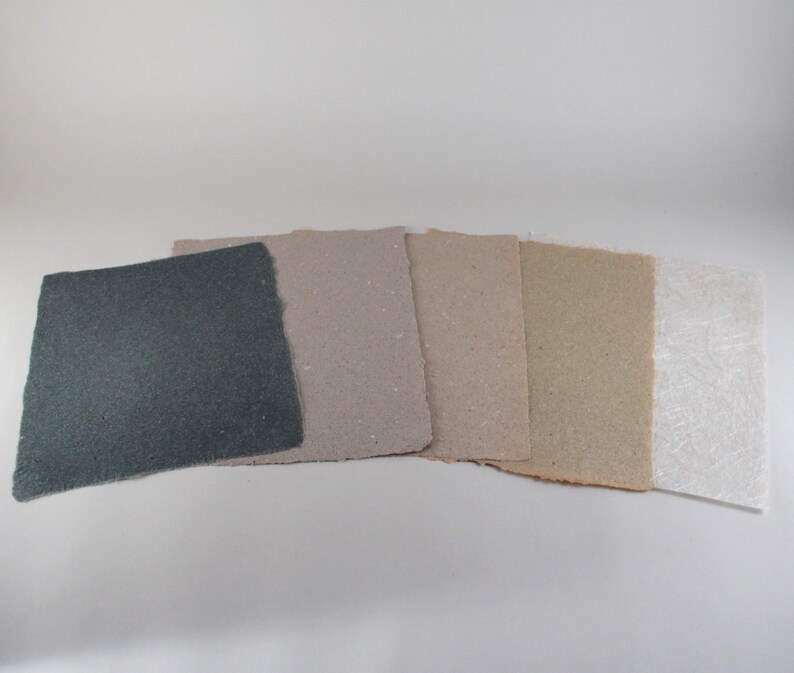May include: A collection of handmade paper sheets in various neutral tones. The sheets range in color from dark gray to off-white, showcasing different textures and finishes. The edges of the paper are rough-cut, adding to the artisanal aesthetic.