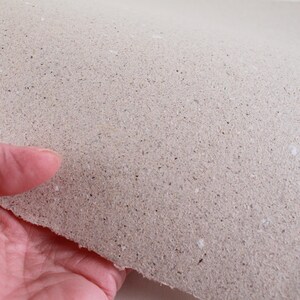 May include: A sheet of handmade paper with a rough, textured surface. The paper is a light beige color with small flecks of darker material throughout. The edge of the paper is uneven, suggesting it was torn by hand.