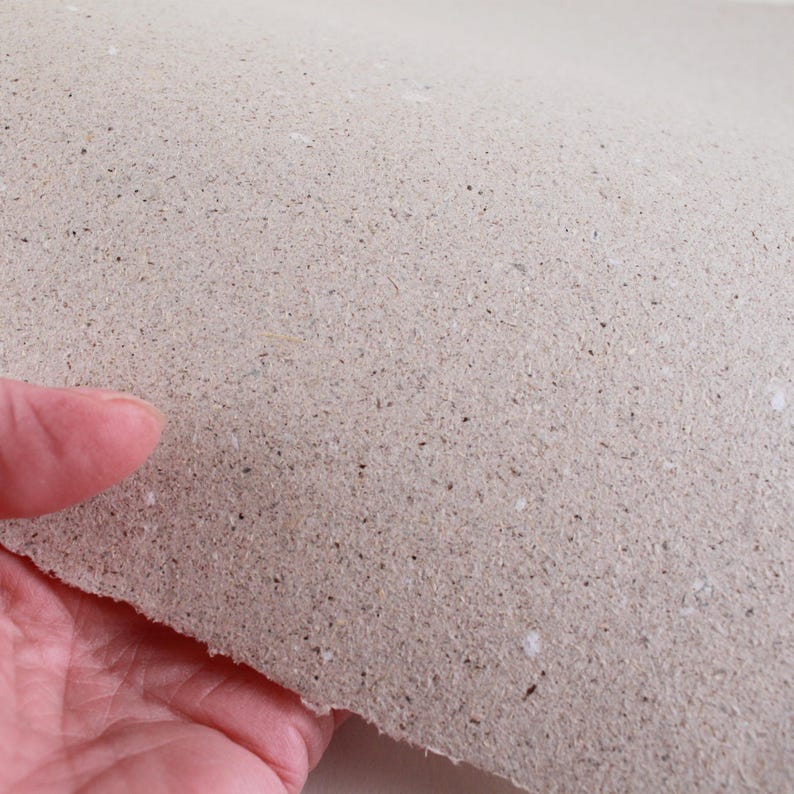 May include: A sheet of handmade paper with a rough, textured surface. The paper is a light beige color with small flecks of darker material throughout. The edge of the paper is uneven, suggesting it was torn by hand.