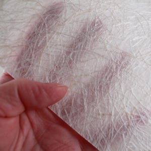 May include: Close-up of a sheet of handmade paper with a textured, fibrous surface. The paper is primarily white with subtle beige and brown fibers interwoven throughout. The paper is held in a hand, with the skin tone visible through the translucent material.