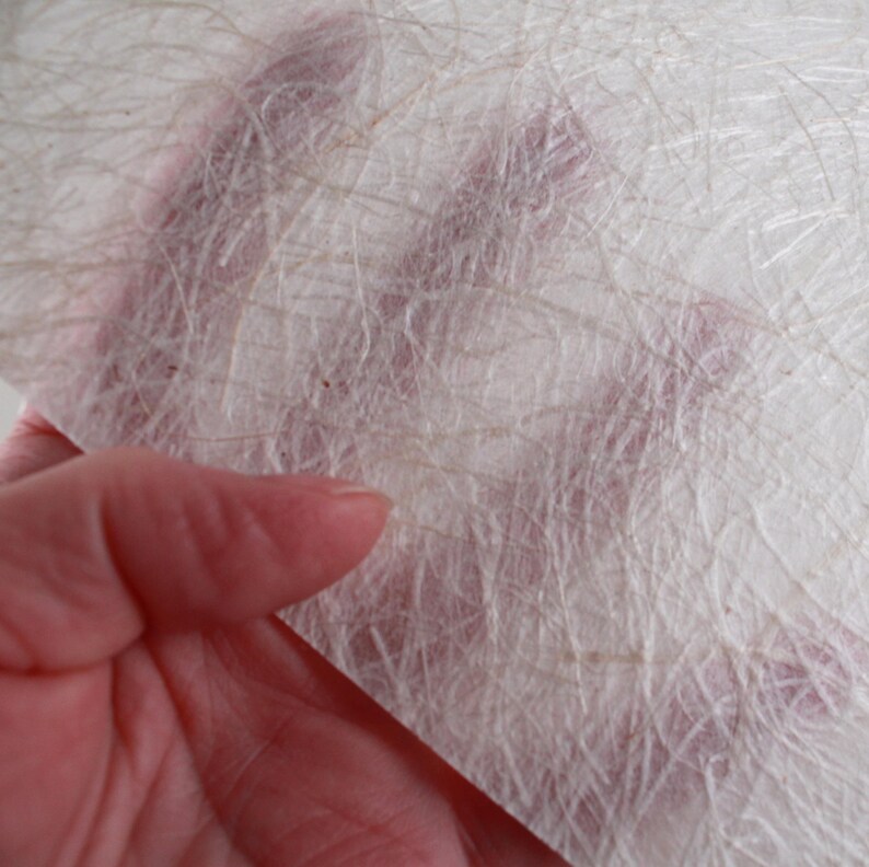 May include: Close-up of a sheet of handmade paper with a textured, fibrous surface. The paper is primarily white with subtle beige and brown fibers interwoven throughout. The paper is held in a hand, with the skin tone visible through the translucent material.