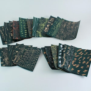 May include: A selection of folded paper cards featuring abstract designs in green, brown, and gold hues. The cards are arranged in a fan shape, displaying diverse patterns. The designs include lines, textures, and organic forms, creating a visually interesting display.