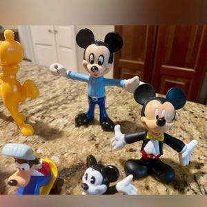 May include: A collection of colorful Disney character figurines, including Mickey Mouse in various outfits and a yellow lion. The Mickey Mouse figures are in blue, black, and red attire. The lion is a bright yellow color.