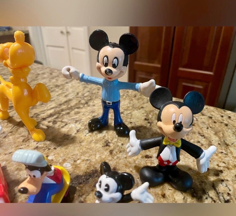 May include: A collection of colorful Disney character figurines, including Mickey Mouse in various outfits and a yellow lion. The Mickey Mouse figures are in blue, black, and red attire. The lion is a bright yellow color.