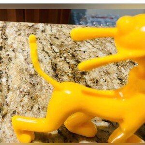 May include: A bright yellow, stylized figurine of a quadruped animal, possibly a lion or dog, in a playful pose. The figurine has a smooth, glossy finish and is set against a speckled, light brown surface.