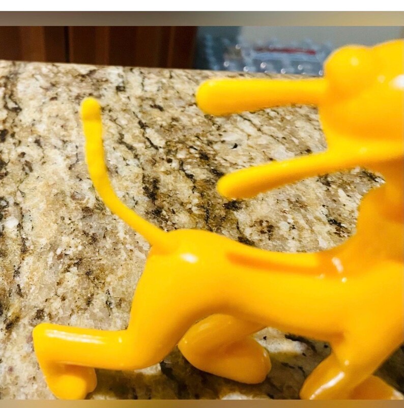 May include: A bright yellow, stylized figurine of a quadruped animal, possibly a lion or dog, in a playful pose. The figurine has a smooth, glossy finish and is set against a speckled, light brown surface.