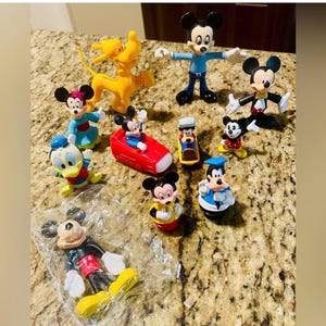 May include: A collection of colorful Mickey Mouse and friends figurines. The figurines include Mickey Mouse, Minnie Mouse, Donald Duck, Pluto, and others. The figures are in various poses and outfits, with bright colors.