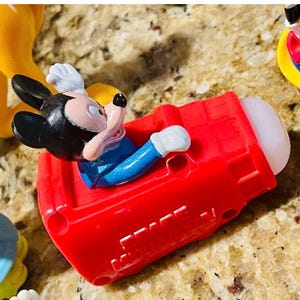 May include: A Mickey Mouse figurine in a red vehicle. Mickey is wearing a blue shirt, white gloves, and black ears. The vehicle is red with a white tip. Other figurines are visible in the background.