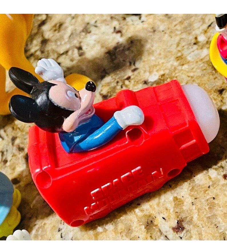 May include: A Mickey Mouse figurine in a red vehicle. Mickey is wearing a blue shirt, white gloves, and black ears. The vehicle is red with a white tip. Other figurines are visible in the background.