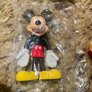 May include: A Mickey Mouse figurine in a clear plastic bag. The figure has a tan face, black ears, a black body, red shorts, white gloves, and yellow shoes. The figurine is a classic Disney character.