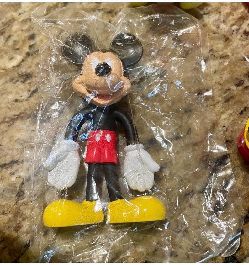 May include: A Mickey Mouse figurine in a clear plastic bag. The figure has a tan face, black ears, a black body, red shorts, white gloves, and yellow shoes. The figurine is a classic Disney character.