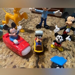 May include: A collection of Disney character figurines, including Mickey Mouse, Pluto, and Goofy. Mickey Mouse is featured in a red car, and Goofy is on a yellow sled. The figurines are in various poses and outfits.