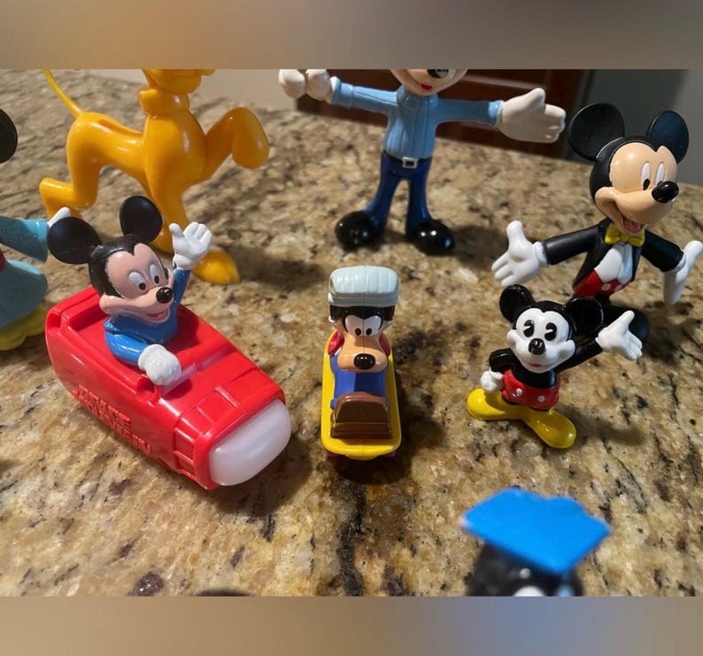May include: A collection of Disney character figurines, including Mickey Mouse, Pluto, and Goofy. Mickey Mouse is featured in a red car, and Goofy is on a yellow sled. The figurines are in various poses and outfits.