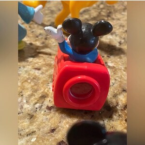 May include: A red toy train car with a black Mickey Mouse figure waving from inside. The figure is wearing a blue shirt and white gloves. Other toy figures are visible in the background.