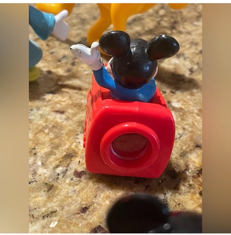 May include: A red toy train car with a black Mickey Mouse figure waving from inside. The figure is wearing a blue shirt and white gloves. Other toy figures are visible in the background.