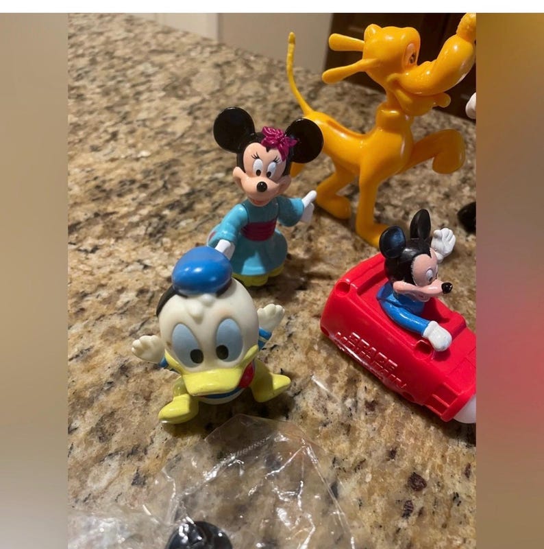 May include: A collection of Disney character figurines, including Mickey Mouse in a red car, Minnie Mouse in a blue dress, Donald Duck, and Pluto. The figures are brightly colored and made of plastic.